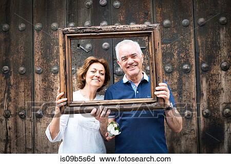 Stock Image - Portrait of senior couple, holding wooden frame in front of their faces, Mexico City, Mexico. Fotosearch