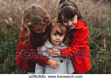 Stock Photograph - Portrait of two girls hugging toddler sister in field. Fotosearch