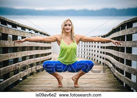 Stock Image - Portrait of young woman crouching in yoga pose on wooden pier. Fotosearch