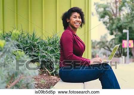 Portrait of young woman sitting on wall, using laptop View Large Photo Image Stock Photography - Portrait of young woman sitting on wall, using laptop. Fotosearch