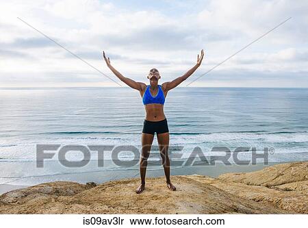 Portrait of young woman standing on rocks beside sea, arms raised, looking up View Large Photo Image Stock Photography - Portrait of young woman standing on rocks beside sea, arms raised, looking up. Fotosearch