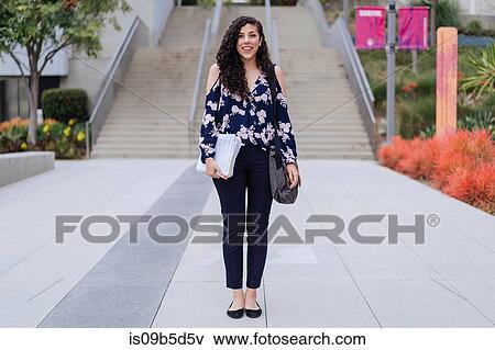 Portrait of young woman standing outdoors, holding bag and paperwork View Large Photo Image Stock Photo - Portrait of young woman standing outdoors, holding bag and paperwork. Fotosearch