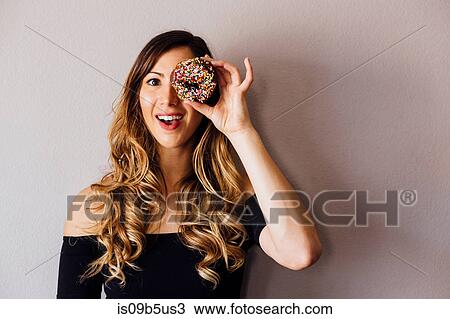 Stock Image - Portrait of young woman with long blond hair holding doughnut hole over eye. Fotosearch
