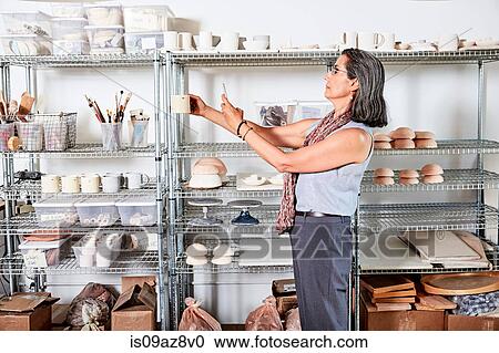 Stock Image - Potter in workshop photographing pottery with smartphone. Fotosearch