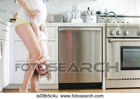 Stock Photograph - Pregnant woman holding baby daughter upside down in kitchen. Fotosearch