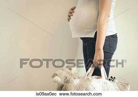 Stock Image - Pregnant woman, holding bag with cuddly toy inside, mid section. Fotosearch