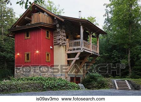 Picture - Rear view of cottage style log home with stone, beige stucco and red vertical wood siding exterior finishes and timber wood balcony at dusk in summer. Fotosearch
