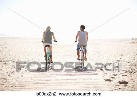 Stock Image - Rear view of couple cycling on boardwalk at Venice Beach, Los Angeles, California, USA. Fotosearch