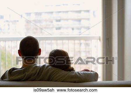 Stock Image - Rear view of couple sitting on sofa looking out of window at apartment building. Fotosearch