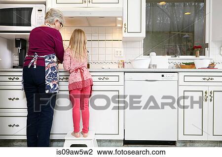 Stock Image - Rear view of girl and grandmother standing at kitchen hob. Fotosearch