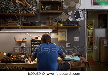 Stock Image - Rear view of guitar maker sitting at desk manufacturing guitar. Fotosearch