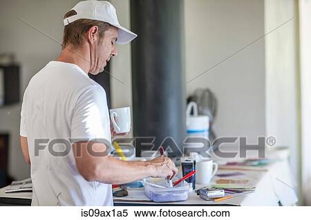 Stock Photography - Rear view of man holding coffee cup mixing paint in plastic container. Fotosearch