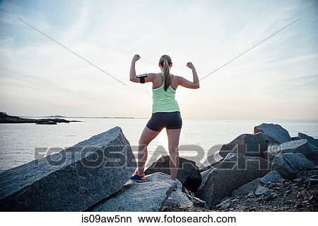 Picture - Rear view of woman standing on rocks arms raised flexing muscles. Fotosearch