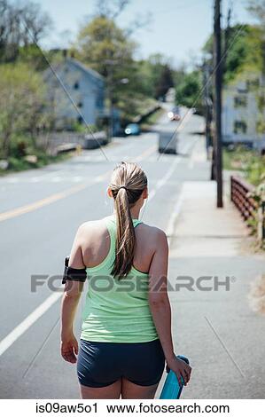Stock Image - Rear view of woman wearing activity tracker carrying water bottle walking along road. Fotosearch