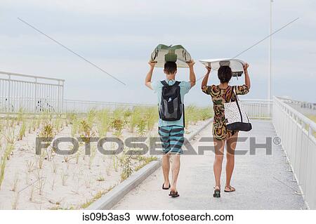 Stock Image - Rear view of young couple carrying surfboards on heads, Rockaway Beach, New York State, USA. Fotosearch