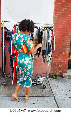 Stock Photo - Rear view of young female fashion blogger with afro hair looking at sidewalk clothes rail, New York, USA. Fotosearch