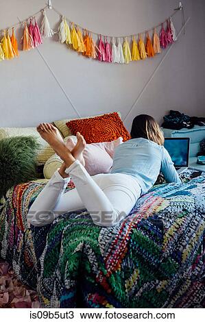 Stock Image - Rear view of young woman lying on shabby chic bed looking at laptop. Fotosearch