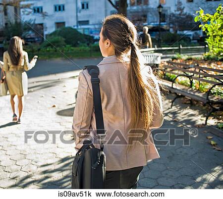 Stock Image - Rear view of young women walking through city park. Fotosearch