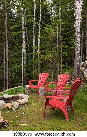 Picture - Red plastic garden chairs around rock fire pit in backyard of cottage-style log home in summer, Quebec, Canada. Fotosearch