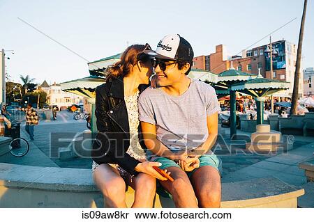 Stock Photography - Romantic young couple sitting on wall at coast, Venice Beach, California, USA. Fotosearch