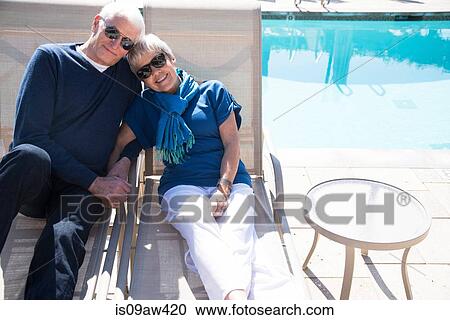 Stock Image - Senior couple relaxing on sun loungers beside pool. Fotosearch