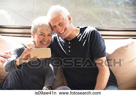 Picture - Senior couple, sitting on sofa, taking self portrait, using smartphone. Fotosearch
