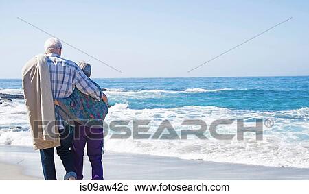 Stock Image - Senior couple standing on beach, looking at ocean, rear view. Fotosearch