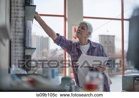 Stock Image - Senior female carpenter reaching for paperwork in furniture making workshop. Fotosearch