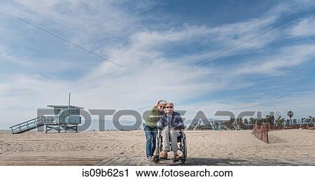 Stock Image - Senior man in wheelchair with wife pointing from beach, Santa Monica, California, USA. Fotosearch