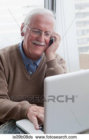 Stock Photograph - Senior man using mobile phone and laptop at home. Fotosearch