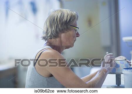Stock Image - Senior woman in pottery workshop, painting bowl. Fotosearch