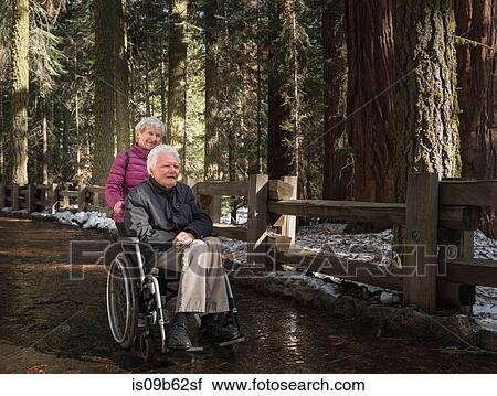 Stock Image - Senior woman pushing husband in wheelchair through forest at Sequoia National Park, California, USA. Fotosearch