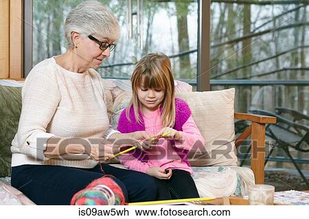 Stock Image - Senior woman teaching granddaughter knitting on living room sofa. Fotosearch