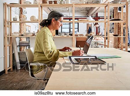 Side view of woman at desk in workshop using laptop View Large Photo Image Stock Photograph - Side view of woman at desk in workshop using laptop. Fotosearch
