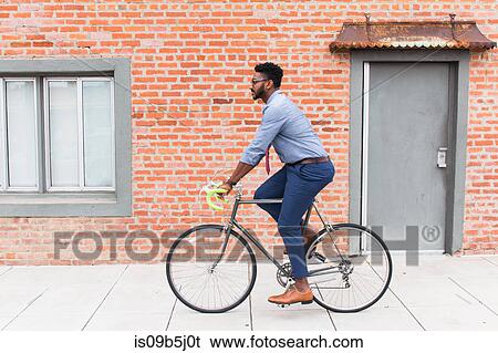 Stock Photograph - Side view of young man cycling along sidewalk. Fotosearch