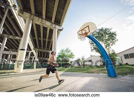 Stock Image - Side view of young man holding basketball running to basketball hoop. Fotosearch