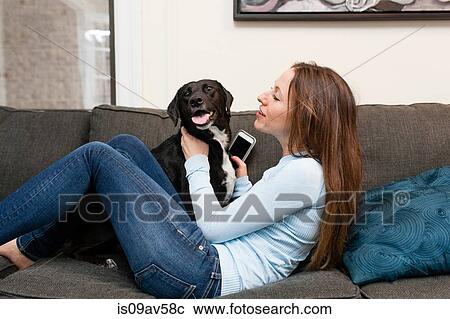 Stock Image - Side view of young woman on sofa holding smartphone playing with dog. Fotosearch