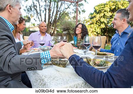 Stock Image - Six mature adult friends holding each others hands at garden party table. Fotosearch