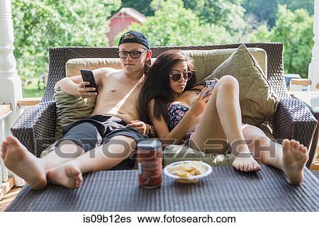 Teenage couple wearing bikini and swim shorts reclining on patio sofa reading smartphones View Large Photo Image Stock Photograph - Teenage couple wearing bikini and swim shorts reclining on patio sofa reading smartphones. Fotosearch