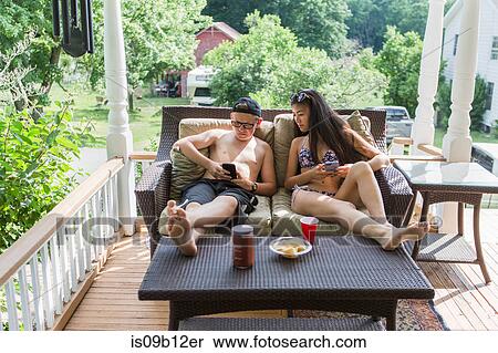 Teenage couple wearing bikini and swim shorts reclining on patio sofa sharing smartphone reading View Large Photo Image Stock Photography - Teenage couple wearing bikini and swim shorts reclining on patio sofa sharing smartphone reading. Fotosearch