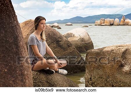 Stock Photo - Teenage girl sitting cross legged on rocks by sea looking away. Fotosearch