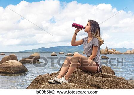 Stock Image - Teenage girl sitting on rocks drinking from water bottle. Fotosearch