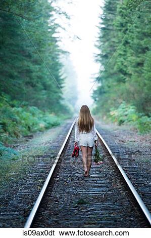 Stock Photograph - Teenage girl, walking along train track, holding red shoes and flowers, rear view. Fotosearch