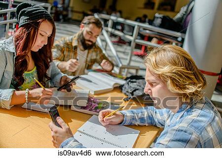 Three adult college students making notes and using smartphones at classroom desk View Large Photo Image Stock Photograph - Three adult college students making notes and using smartphones at classroom desk. Fotosearch