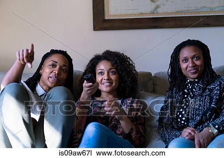 Stock Image - Three female friends, sitting on sofa, watching TV. Fotosearch