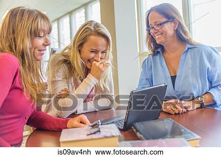 Stock Image - Three women using laptop and giggling at desk in library. Fotosearch