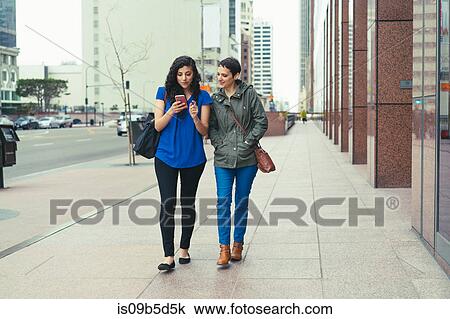 Stock Image - Two female friends walking along street, looking at smartphone. Fotosearch