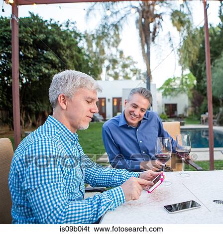 Stock Photograph - Two mature men looking at smartphone touchscreen at garden party table. Fotosearch