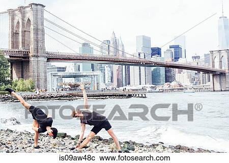 Stock Image - Two men doing yoga on riverside in front of Brooklyn Bridge, New York, USA. Fotosearch