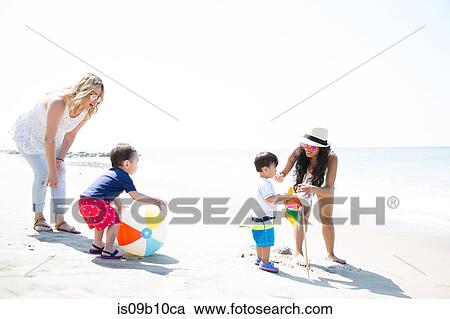 Stock Image - Two mothers and baby sons playing with beach ball and pinwheel on beach, Malibu, California, USA. Fotosearch